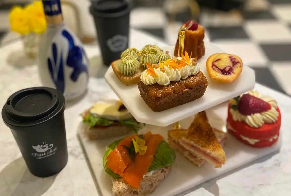 Torre de dos pisos sobre mesa de mármol: sándwiches, tartaletas, cannelé, budín y red velvet, junto a vasos de Petit Chat