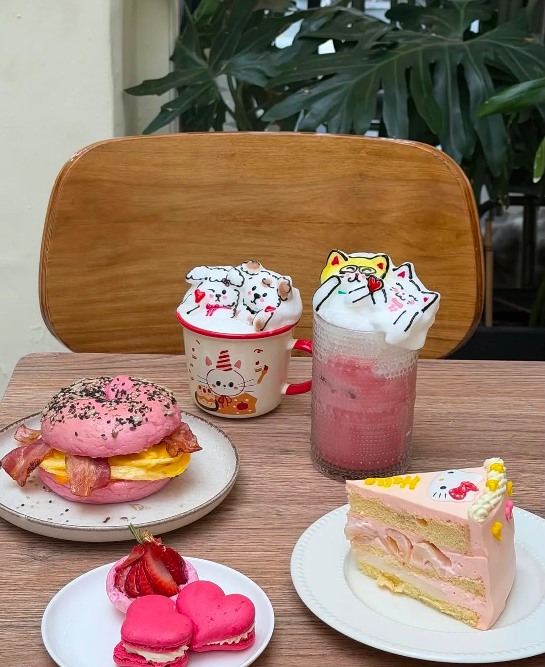 Mesa en tonos rosa de Petit Chat: bagel relleno con huevo y bacon, tazas con arte latte de gatitos, frappé rosa y porción de torta Hello Kitty con macarons con forma de corazón